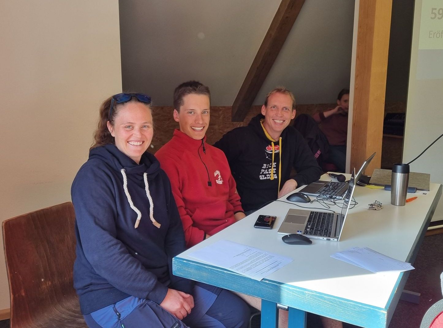 Drei Personen sitzen hinter einem Tisch und lächeln in die Kamera.