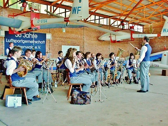 Eine Blaskapelle spielt in einem Hangar unter aufgehängten Segelflugzeugen.