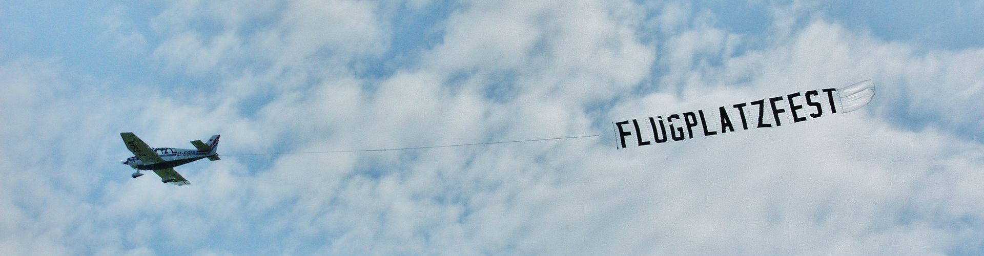 Ein Motorflugzeug vor leicht bewölktem Himmel. Es schleppt ein Banner auf dem steht: