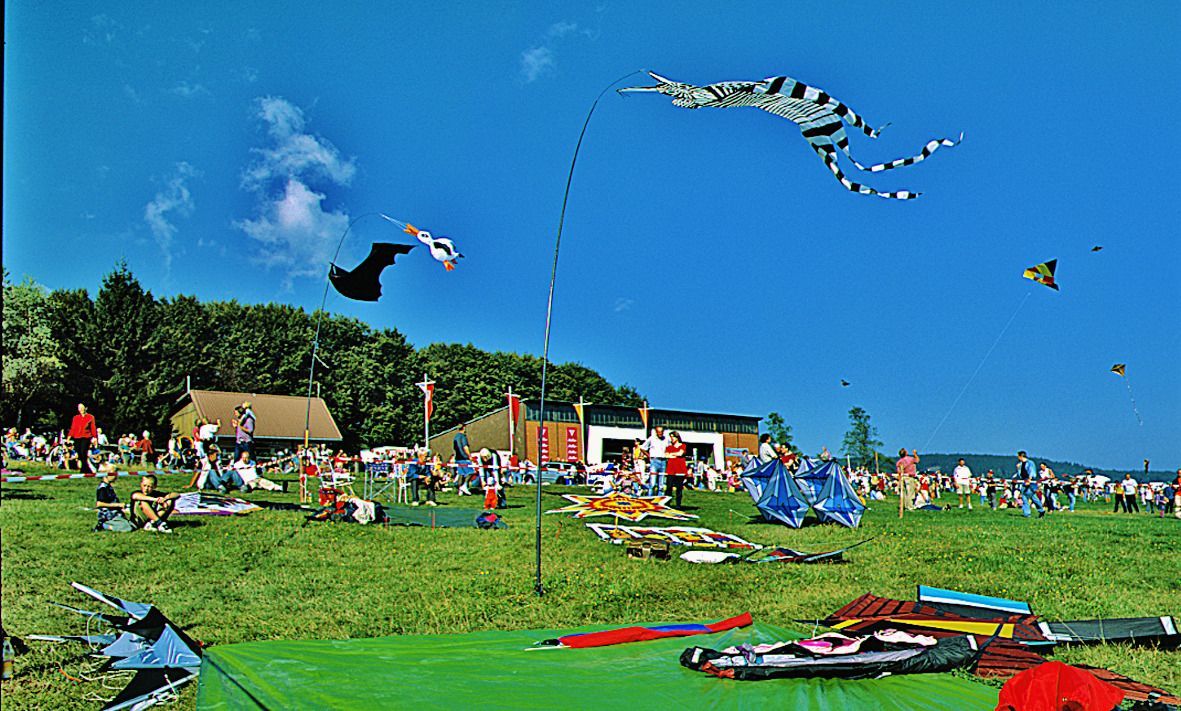 Der Flugplatz Hütten/Hotzenwald bei einem Drachenfest. Im Hintergrund die Halle und die Fliegerklause, im Vordergrund viele Leute und bunte Drachen vor blauem Himmel.