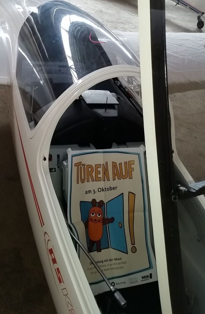 Eine Flugzeughalle. Eine ASK 21 mit geöffneter Haube. Auf dem Sitz liegt ein Plakat der Sendung mit der Maus. Aufschrift: Türen auf am 3. Oktober.