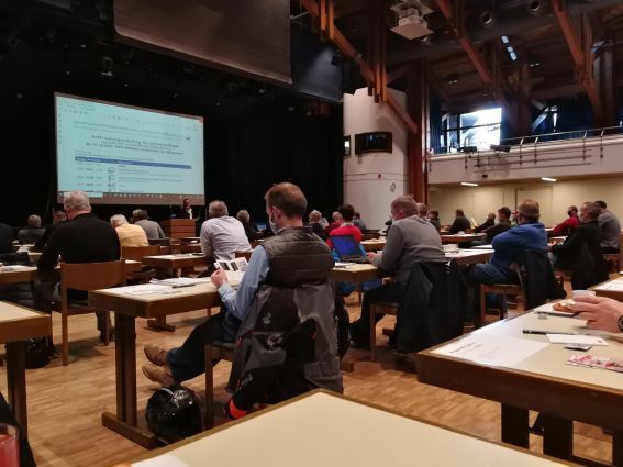 Ein großer Saal mit Leinwand. die Zuhörer sitzen mit größerem Abstand an Einzeltischen.