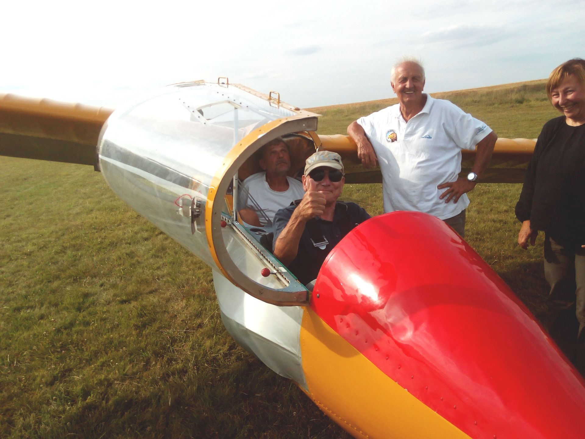 Ein gelb-orange lackiertes, doppelsitziges Segelflugzeug mit offener Haube, beide Sitze besetzt und zwei Personen stehen daneben.